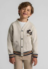 Šypsosi berniukas su šviesiai rudais plaukais, vilkintis kreminį švarką su „Los Andželas“ pledu, baltą marškinėlį ir moliūgų spalvos kelnes prieš paprastą foną.