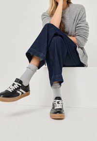 Osoba sedící se zkříženýma nohama, oblečená ve šedém svetru, tmavě modrých širokých kalhotách, šedých ponožkách a černých teniskách s bílými tkaničkami.