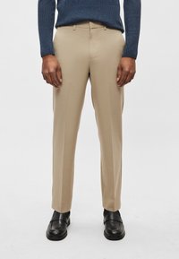 Lehúzott, bézs színű, könnyű anyagból készült nadrág, egyenes szabással és oldalzsebekkel, fekete bőrcipővel párosítva.