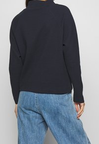 Marinblå strukturerad tröja med hög krage och raglanärmar, i kombination med ljusblå denimbyxor. Slät tygfinish.