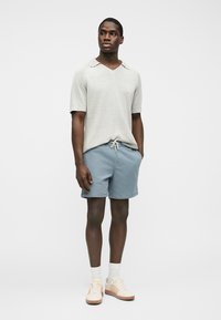 Homme noir debout portant un polo en maille gris clair à manches courtes, un short bleu à cordon de serrage, des chaussettes blanches et des baskets crème.