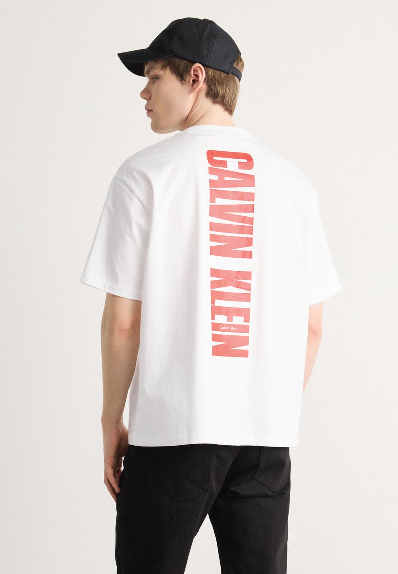 Hvit bomulls T-skjorte med en vertikal rød "CALVIN KLEIN" logo på ryggen. Korte ermer, avslappet passform, kombinert med svarte bukser og en caps.
