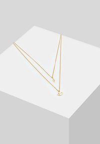 Collier doré à deux chaînes. Une chaîne présente un pendentif en forme d'éclair, tandis que l'autre affiche un motif d'œil avec une pierre.