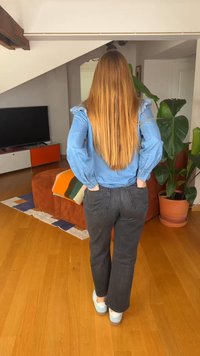 Persona con lunghi capelli biondi che indossa una camicia blu e pantaloni neri, in piedi con le mani nelle tasche posteriori in un soggiorno.