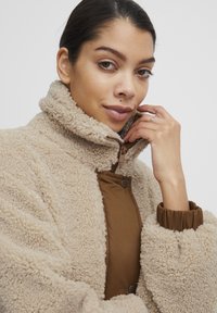 Beige fleecejacka med hög krage, tryckknappsstängning och kontrasterande brun innerfoder. Har elastiska ärmslut och texturerad tyg.
