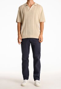 Beige kortärmad polo med V-ringning, med vertikala ribbade detaljer. Matchad med marinblå chinos och vita sneakers.