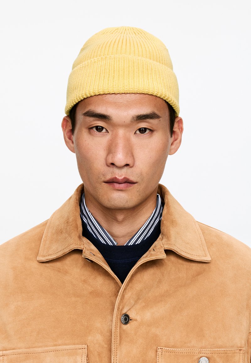 Gorro amarillo de punto acanalado, chaqueta de ante color coñac, camisa a rayas azules y blancas. Primer plano del rostro con expresión neutra sobre un fondo liso.