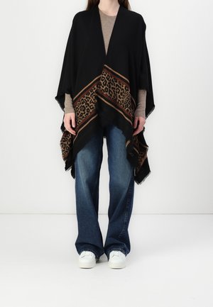 Personne portant un châle noir avec une bordure imprimée léopard marron sur un pull beige, un jean bleu à jambes larges et des baskets blanches, debout contre un mur blanc.