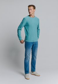 Lichtblauw langemouwen shirt van textuurstof, ronde halslijn en geribde manchetten, gedragen met blauwe jeans en witte sneakers.