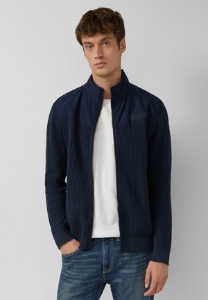 Navy zip-up jacket with a textured knit pattern, ribbed cuffs, and a high collar. Paired with a white shirt and blue jeans.