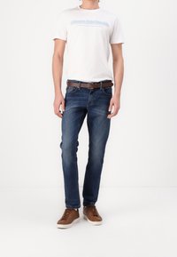 Jeans blu scuro con una vestibilità slim, una cintura marrone e sneaker beige. Il modello indossa una maglietta bianca con testo blu in una posa casual.