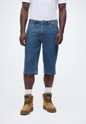 Mężczyzna w niebieskich dżinsowych szortach za kolano, białych skarpetkach, beżowych butach sznurowanych i białej koszuli z krótkim rękawem stojący na jednolitym tle.