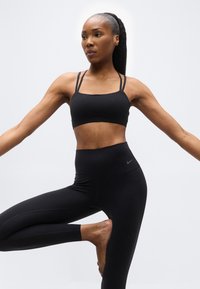 Γυναίκα με μαύρα αθλητικά ρούχα ισορροπώντας στο ένα πόδι με τα χέρια εκτεταμένα πάνω σε λευκό φόντο.