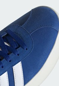 Blå mockaskor med vita läderränder, strukturerad yta, rund tå och blå skosnören mot en vit gummisula.