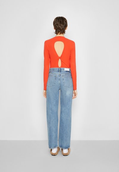 Camisola vermelha de mangas longas com abertura nas costas, combinada com jeans de perna larga em azul claro de cintura alta e sapatos castanhos de slip-on.