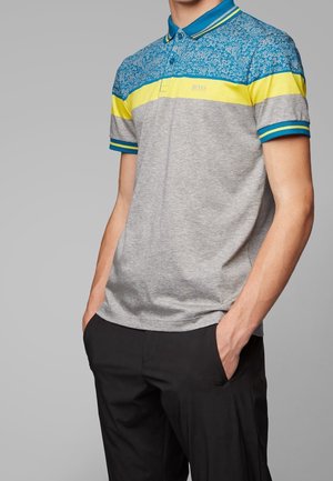 Homme portant un polo à manches courtes avec des blocs de couleur bleu floral, jaune et gris, associé à un pantalon noir, les mains dans les poches.