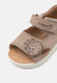 Beige sandaalit, jotka on valmistettu pehmeästä kirjoittamattomasta nahasta, suljetulla varvaskappaleella, säädettävällä hihnalla ja pyöreällä kirahvidetaljilla hihnassa. Valkoinen kumipohja.