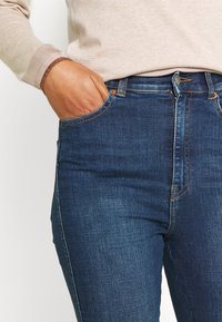 Persona vestida con jeans de mezclilla azules y un suéter beige, con la mano derecha en el bolsillo delantero de los jeans.