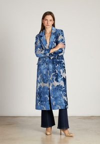 Abrigo de patrón floral azul con cuello chal, hecho de un tejido texturizado, que incluye un cinturón y dos bolsillos delanteros, combinado con jeans de pierna ancha.