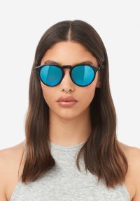 Černé sluneční brýle s kulatými, modrými zrcadlovými čočkami. Tenké obruba s unikátním designem nosníku. Nosené osobou se středně dlouhými, rovnými vlasy.