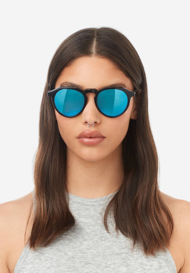Černé sluneční brýle s kulatými, modrými zrcadlovými čočkami. Tenké obruba s unikátním designem nosníku. Nosené osobou se středně dlouhými, rovnými vlasy.