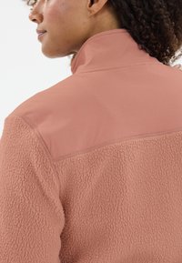 Fleecejacke in sanftem Rosa, mit einem strukturierten unteren Bereich und einer glatten oberen Sektion. Hoher Kragen ohne sichtbare Verschlüsse.