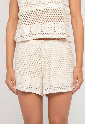 Femme portant un short en crochet couleur crème et un haut sans manches assorti avec des motifs circulaires et une taille à cordon.