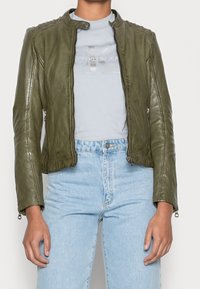 Olivgrön läderjacka med en figurnära design, hög krage och axeldetaljer. Dragkedjeslutar, stylad med ljusblå skjorta och jeans.