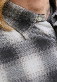 Camisa a cuadros con patrones en gris y negro. La tela es suave y con una textura sutil, complementada por un botón blanco en el cuello.
