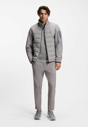Szara kurtka puchowa z wysokim kołnierzem i zamkiem z przodu, noszona na szarym topie, zestawiona z szarymi zwężanymi spodniami i trampkami.