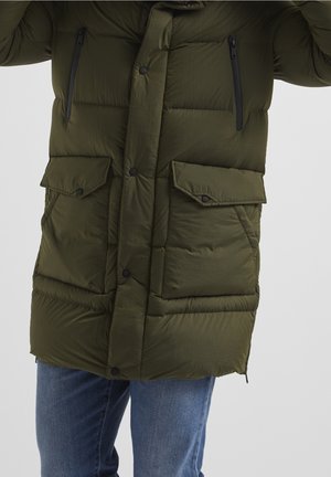 Lunga giacca imbottita verde oliva con quattro tasche frontali e bottoni a pressione neri, indossata con jeans blu su sfondo chiaro.