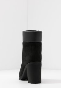 Svart ankelboot i mocka och syntetiska material, med en kraftig klack, rund tå och logotypdetalj på sidan.