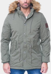Parka verde oliva con cappuccio foderato in pelliccia sintetica, bottoni frontali, cordini e tasche laterali. Realizzata in un tessuto testurizzato.