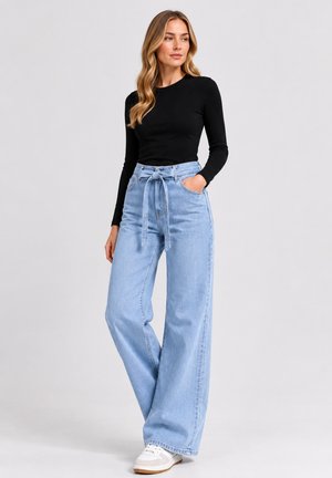 Femme portant un haut noir à manches longues, un jean large bleu clair taille haute avec ceinture à nouer, et des baskets blanches, debout devant un fond uni.