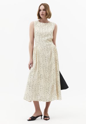 Vrouw draagt een mouwloze crèmekleurige midi-jurk met zwarte polkadots, zwarte open-toe hakken en houdt een zwarte gestructureerde tas vast.