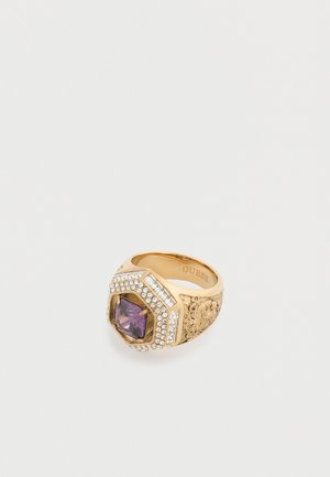 Bague en or avec des gravures florales complexes, pierre précieuse centrale violette et petits cristaux clairs autour, gravée "GUESS" à l'intérieur de l'anneau.