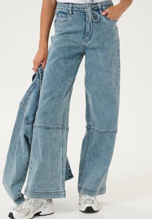 Helles blau, weit geschnittene Denim-Jeans mit einem Kordelzugbund, kontrastierenden Nähten und einer entspannten Passform. Kombiniert mit weißen Sneakers.