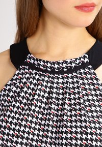 Top con estampado de pata de gallo en blanco y negro con acentos rojos, presenta un escote redondo negro y detalle de tela fruncida.