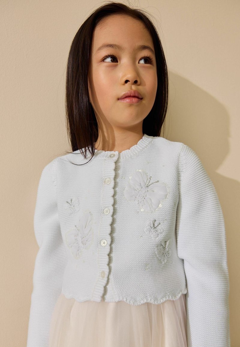 Jeune fille aux longs cheveux foncés portant un cardigan blanc tricoté décoré de motifs floraux, regardant sur le côté sur un fond neutre.