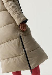 Béžový puffr kabát s prešívaným dizajnom, má bočný zips a priliehavé manžety. Kabát má lesklý povrch a voľný strih.