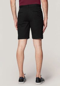 Czarne bawełniane szorty o dopasowanym kroju, z dwiema kieszeniami z tyłu, zestawione z czarnymi trampkami na nagich nogach.