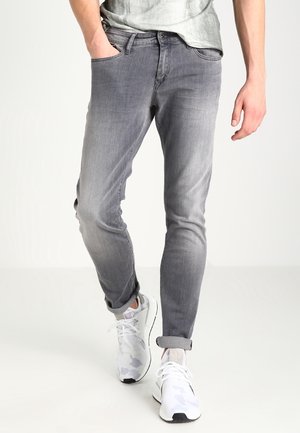 Mężczyzna noszący dopasowane, wyblakłe szare dżinsy z podwiniętymi nogawkami i białe sportowe trampki, stoi z jedną ręką w kieszeni.