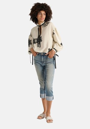 Blusa crema con ricami floreali neri, scollo a strascico e maniche a sbuffo; abbinata a jeans cropped blu chiaro con effetti di usura e sandali beige.