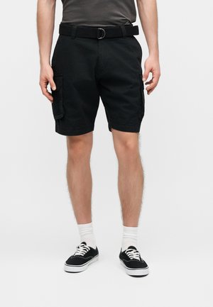 Uomo che indossa pantaloncini cargo neri con cintura, calzini bianchi corti e sneakers nere basse, in piedi su uno sfondo bianco semplice.