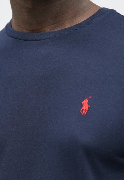 Tmavomodré bavlnené tričko s okrúhlym výstrihom, na ľavej strane malé červené vyšívané logo pólistu.