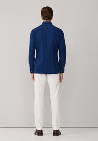 Camicia a maniche lunghe blu navy con colletto con bottoni, realizzata in tessuto liscio. Indossata con pantaloni bianchi e scarpe scure, vista da dietro.