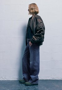 Maslinasto zelena bomber jakna sa satenskim završetkom, slojevito preko crne majice, uparena sa širokim trapericama i obućom sa zelenim potplatom.