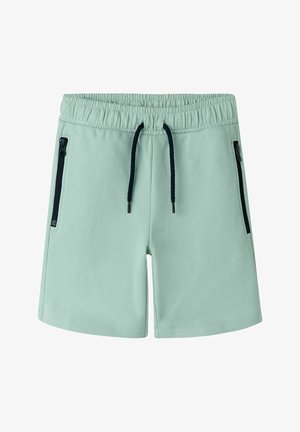 Jasnozielone szorty z elastycznym pasem, czarnym sznurkiem i czarnymi kieszeniami na zamek po bokach, ułożone na białym tle.