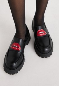 Black leather loafers with a chunky sole, featuring a red lip design on the strap, detailed with a small silver ring accent.
