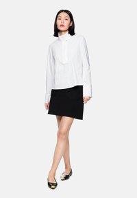 White long-sleeve shirt with a stand collar, paired with a black textured skirt featuring two front pockets, and black flats with a gold toe accent.